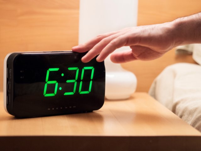 digital alarm clock on a nightstand with a hand touching the top of it