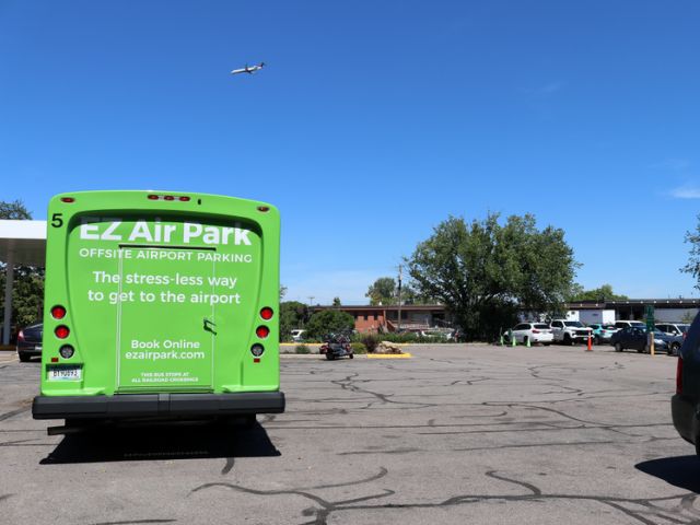 green airport parking shuttle bus in a parking lot with an airplane flying overhead
